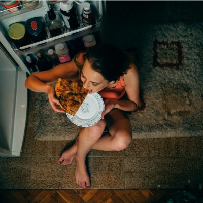 Frau isst kalorienreiches Essen vor dem Kühlschrank um plötzlichen Hunger zu stillen